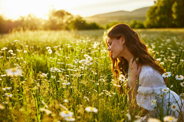 portrait of a beautiful girl, in a light summer dress, sitting in a chamomile field and admiring the flowers