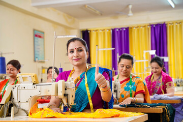 Indian woman showing thumps up while working on sewing machine at textile factory.