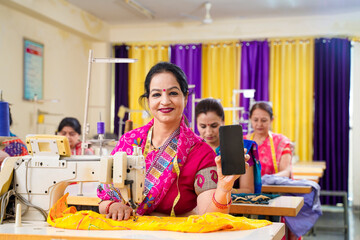 Indian women showing smartphone screen while working on sewing machine at factory