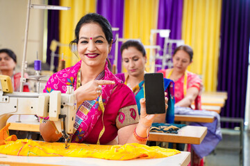 Indian women showing smartphone screen while working on sewing machine at factory