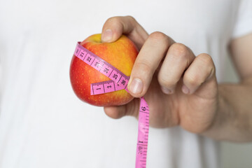 A man in a white T-shirt holds in his hand a red apple with a measuring ribbon close-up. The concept of proper nutrition and healthy lifestyle