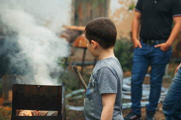 Child boy playing with smoking wooden stick outdoors. Child watching the fire. Barbecue in nature, group of preparing sausages on fire. Group of young friends camping and burning a wood fire.
