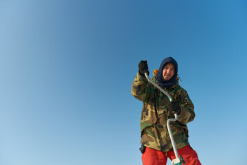 A warmly dressed Asian guy drills a hole in the ice on winter fi