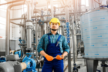 Young worker of chemical factory poses against background of metal structures and stainless steel tanks. Man holds wrench in his hands.