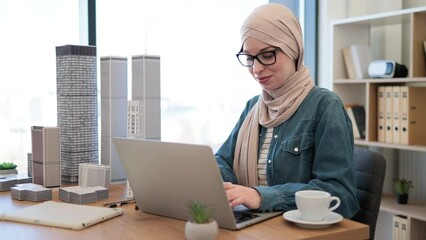 Joyful muslim female wearing hijab and glasses typing on laptop while holding architectural model from office desk indoors. Proficient urban planner developing land use programs of metropolitan area. - Powered by Adobe
