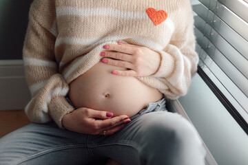Cropped photo of young pregnant woman wearing sweater with application of heart, sitting on floor,...