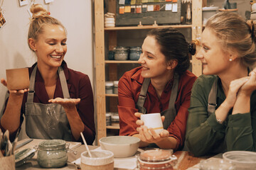 A company of three pretty young women friends make ceramic mugs in a pottery workshop.