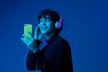 Man smile with phone in hand taking selfies with headphones listening to music, portrait dark blue background, neon light, style and trends, mixed light, men's fashion, copy spot