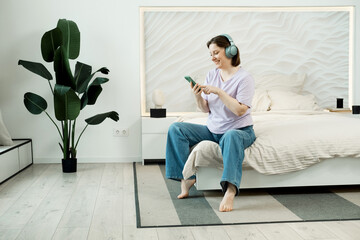 Smiling adult woman sitting on the bed in headphones and listens to music