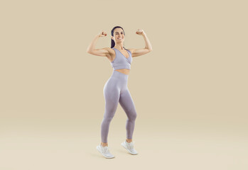 Fototapeta na wymiar Beautiful sporty woman showing off athletic body and muscular arms on beige background. Full length portrait of fitness woman in sportswear showing biceps on camera. Workout sport motivation concept.