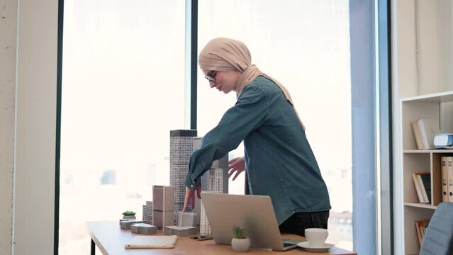 Charming arabian female in traditional hijab working on modern laptop while touching scale model on writing desk in office. Architectural project manager designing plan of modern construction.