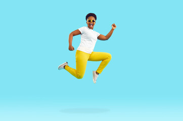 Full body length side profile view portrait happy excited Afro American woman in white tee, yellow pants and cool sunglasses jumping, running and flying high up in air on bright blue studio background