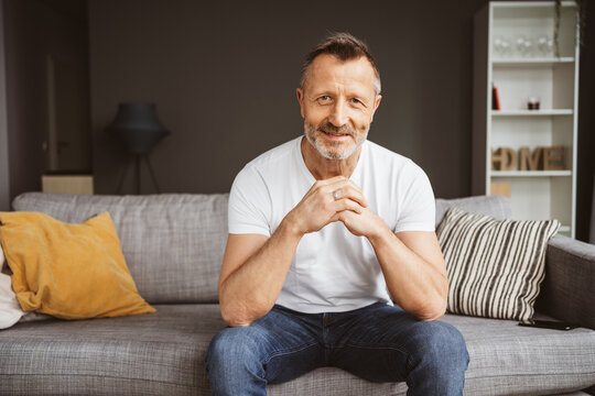 A mature adult man is happily relaxing on a comfortable sofa in his living room, smiling contentedly as he enjoys life at home.