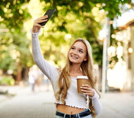 Young teeanager girl walking on a town street and taking selfie while holding takeaway paper coffee cup in her hand - Smiling student using her smartphone with headphones on her neck