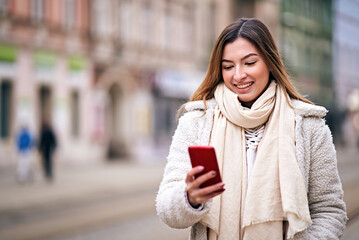Fototapeta premium Cheerful young adult woman walking on city street in cold weather and typing a message on her mobile phone - Millennial girl sharing a story on social media platform