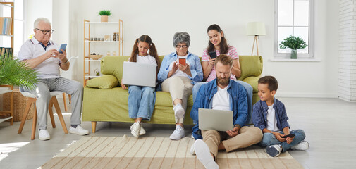 Big family watching gadgets while sitting on sofa at home. Grandparents, dad, mom, daughter and son using digital devices, everyone using their own gadget. Social networks dependence concept