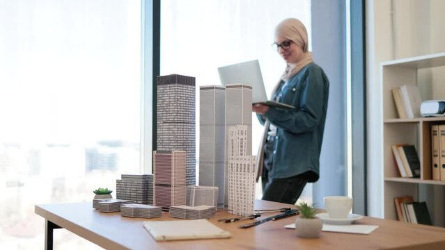 Arabian businesswoman wearing hijab and glasses standing with laptop in hands near panoramic window in creative workplace. Young architect carrying out city complex construction plan using gadgets.