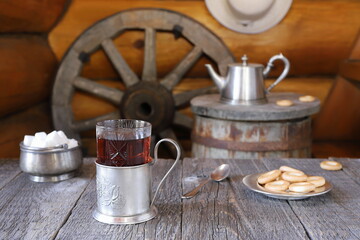 On wooden table  there is a glass of tea in holder next to plate with bagels and other  utensils on background of old wooden barrel and wagon wheel standing near log wall with hanging white hat on it.