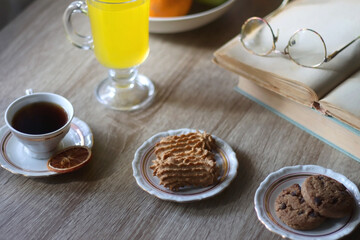 Cup of tea, plates with cookies, glass of orange juice, books, reading glasses, bowl of fruit and candles on the table. Selective focus.