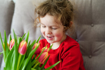 Obraz na płótnie Canvas Adorable smiling little girl in red cloth holding tulips at home