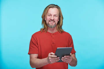 Portrait of mature man in red shirt smiling at camera on blue background and using digital tablet