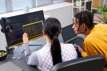 High angle view of two young people writing code in office and pointing at computer screen