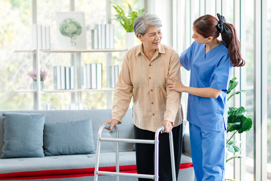 Asian young professional successful friendly female nurse in blue hospital uniform helping supporting physical therapy senior old pensioner unhealthy injury woman patient walking via four legs walker