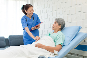 Fototapeta premium Asian professional successful female internship nurse in blue uniform with stethoscope visiting old senior elderly pensioner woman patient laying lying down on bed writing symptoms down on clipboard