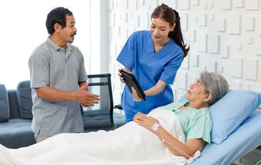 Asian professional successful female assistant internship nurse in blue uniform holding tablet computer visiting discussing showing diagnosis information to old senior unhealthy patient in ward