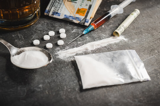 Drugs white pill, syringe with a dose of narcotic substances, white powder cocaine, alcohol drink in a glass and US dollar currency on dark background. Concept of addiction, abuse and bad habits