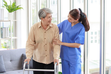 Asian young professional successful friendly female nurse in blue hospital uniform helping supporting physical therapy senior old pensioner unhealthy injury woman patient walking via four legs walker