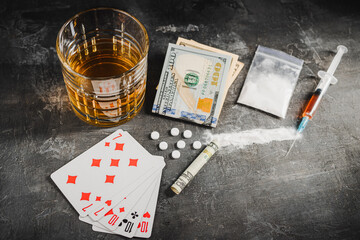 Alcohol drink in a glass, playing cards for poker game, syringe with a dose of drugs, white pills, powder narcotics and US dollar currency on dark background. Concept of addiction, gambling and abuse