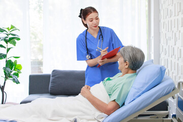 Obraz premium Asian professional successful female internship nurse in blue uniform with stethoscope visiting old senior elderly pensioner woman patient laying lying down on bed writing symptoms down on clipboard