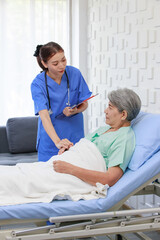 Obraz premium Asian professional successful female internship nurse in blue uniform with stethoscope visiting old senior elderly pensioner woman patient laying lying down on bed writing symptoms down on clipboard