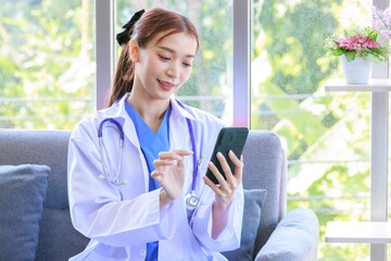 Asian professional successful female doctor in white lab coat with stethoscope sitting smiling using mobile phone posing in hospital office hall way.