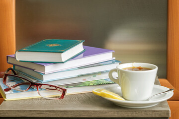 On the wooden surface of the table are a stack of books for reading, women's eyeglasses, a cup of...