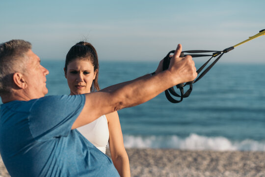 Close-up image of an older overweight white-haired gentleman performing suspension training on the beach under the supervision of his young personal trainer.