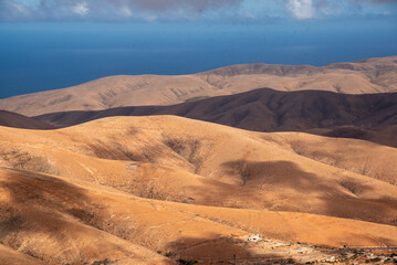 Fototapeta na wymiar Vista panorámica de un impresionante paisaje volcánico y desértico, con grandes montañas rocosas y una pequeña Casa Blanca en una esquina, iluminada por la luz del sol en Fuerteventura, Islas Canaria