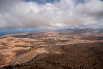 
Vista panorámica de un impresionante paisaje volcánico y desértico, con grandes montañas rocosas y tierra marrón clara, al fondo el mar Fuerteventura, Islas Canarias durante un día nublado.