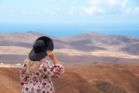 Mujer rubia con un vestido floreado sosteniendo un sombrero de paja negro en medio de un paisaje desértico y volcánico en Fuerteventura en el Mirador del las Peñas, frente a las montañas de las Islas 