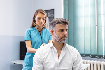 Fototapeta premium A female doctor at the clinic performs auscultation of the lungs of a patient with symptoms of coronavirus or pneumonia. He is coughing the doctor listens to the wheezing in the lungs