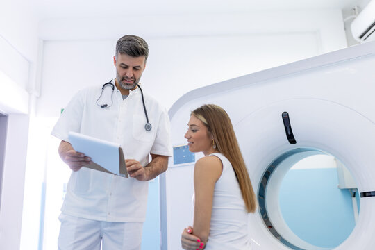 Young medical technician talking to his patient sitting on the CT scanner bed.