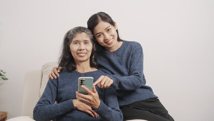 Using mobile, video call, Mother's day concept, young adult female daughter congratulate excited asian elderly mother at sofa with birthday anniversary, two generations family photo, real people.