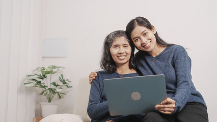 Using laptop, online shopping, Mother's day concept, young adult female daughter congratulate excited asian elderly mother at sofa with birthday anniversary, two generations family photo, real people.