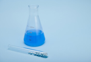 Still life graduated flat bottomed laboratory flask with fluid chemical solution and pharmaceutical therapeutic granules inside a test tube, isolated on blue background. Clinical research and pharmacy