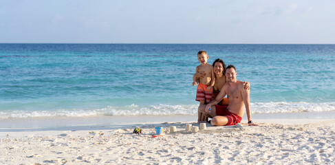 A family of three builds a sand castle on a beach in the Maldives. Family beach holiday concept. Banner