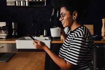African woman using smartphone and drinking coffee while sitting in kitchen