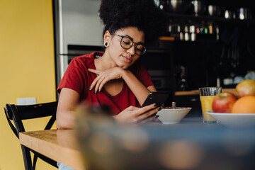 African woman using smartphone while having breakfast in kitchen