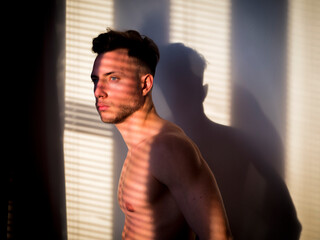 Fototapeta na wymiar Pensive topless man in shadow stripes from window