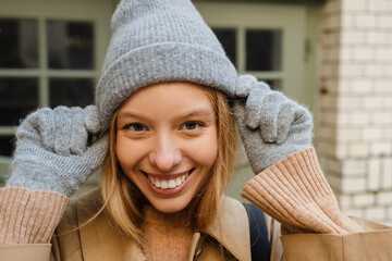 Portrait of woman adjusting warm hat and smiling at camera while standing outdoors
